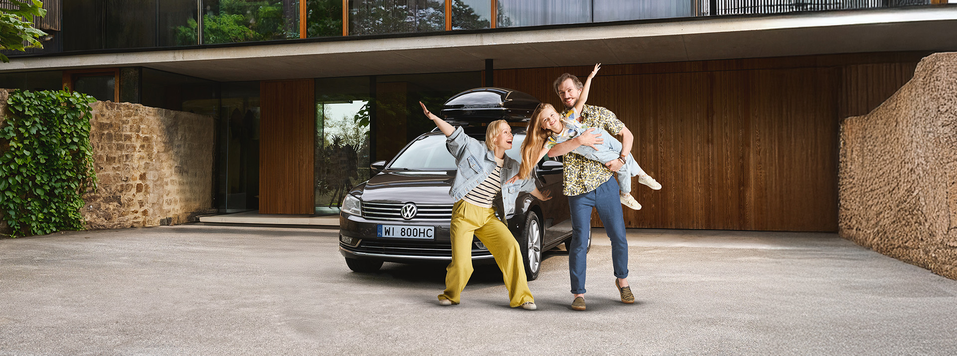 Uśmiechnięta rodzina – kobieta, mężczyzna i dziewczynka – stoi przed czarnym samochodem marki Volkswagen z boxem dachowym, zaparkowanym przed nowoczesnym domem. Mężczyzna trzyma dziecko na rękach w zabawnej pozie, wszyscy wyglądają na gotowych do wyjazdu.