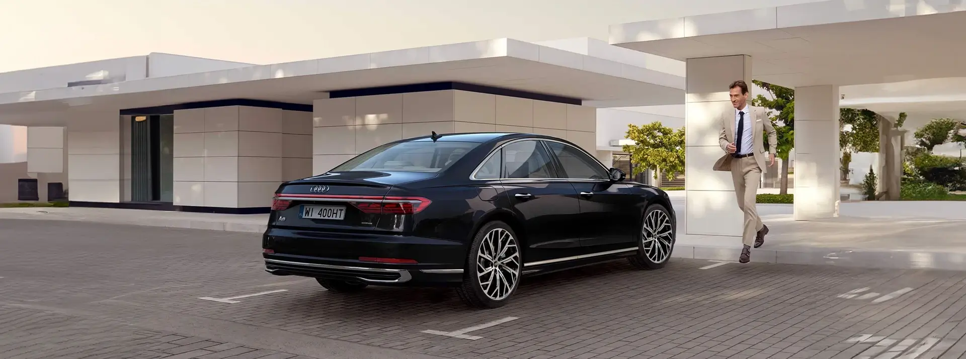 parking przed niskimi budynkami, elegancki mężczyzna podchodzi do czarnej limuzyny Audi A8