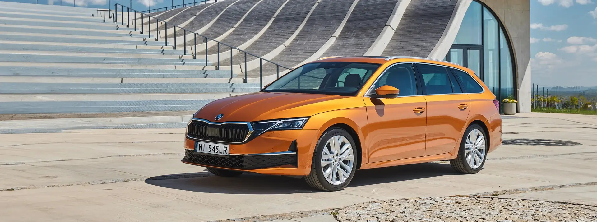 pomarańczowa Skoda Octavia Combi na tle schodów prowadzących do nowoczesnego budynku