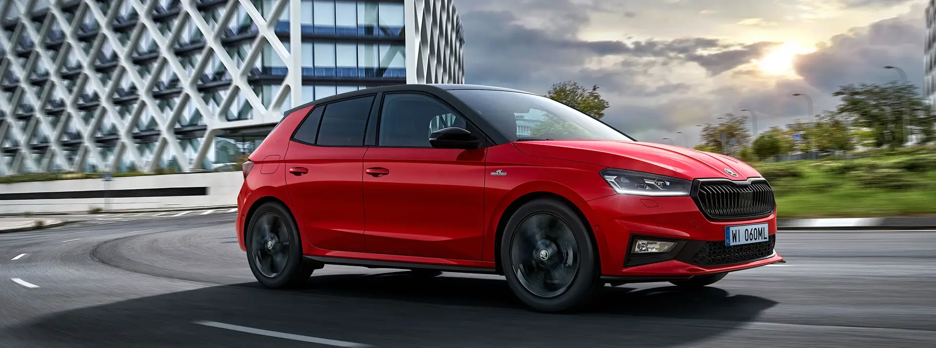 czerwona Skoda Fabia jedzie drogą położoną między miastem a wiejskimi równinami