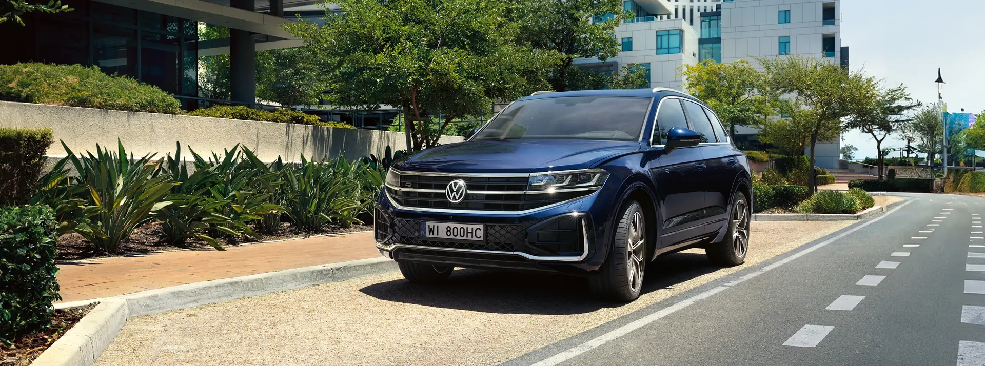 granatowy SUV - Volkswagen Touareg zaparkowany przy ulicy na tle drzew, krzewów i jasnego budynku