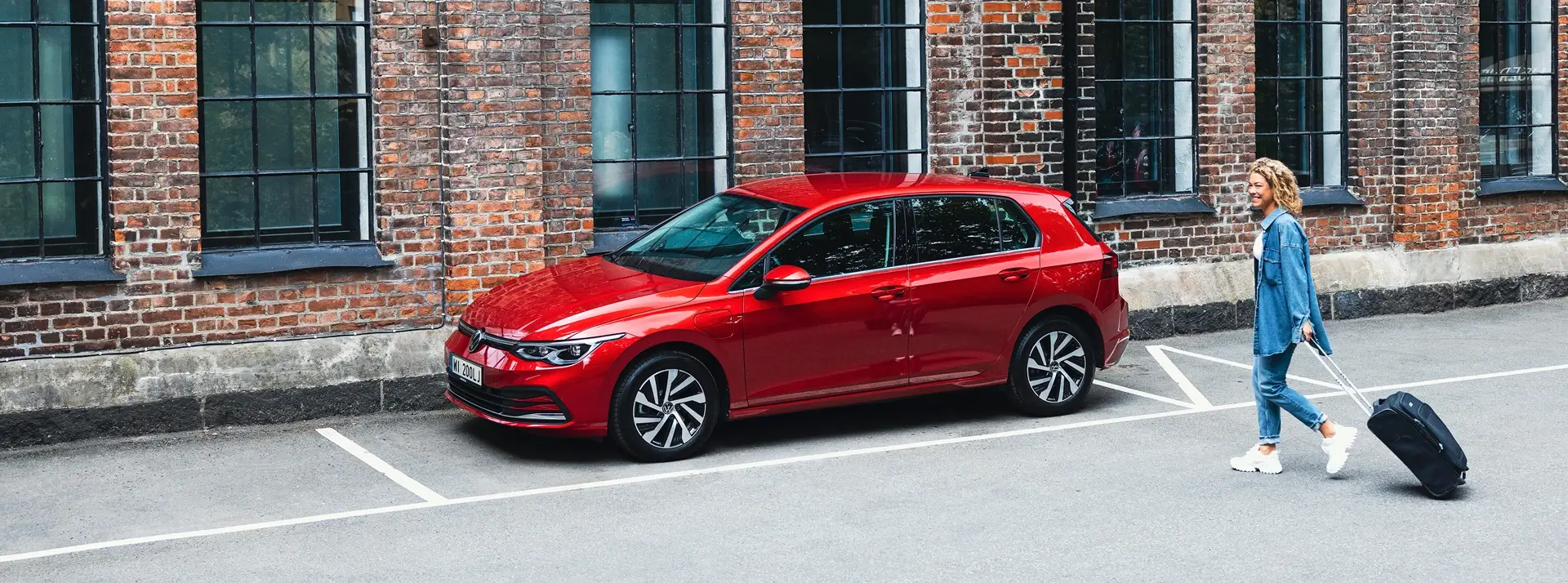 kobieta z walizką idzie w stronę czerwonego Volkswagena Polo zaparkowanego pod budynkiem z czerwonej cegły