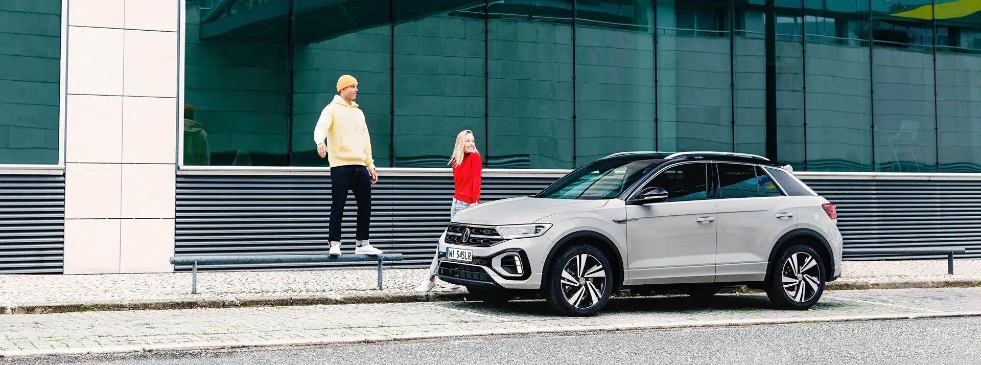 kobieta i mężczyzna obok białego Volkswagena T-Cross, auto jest zaparkowane przed oszklonym budynkiem