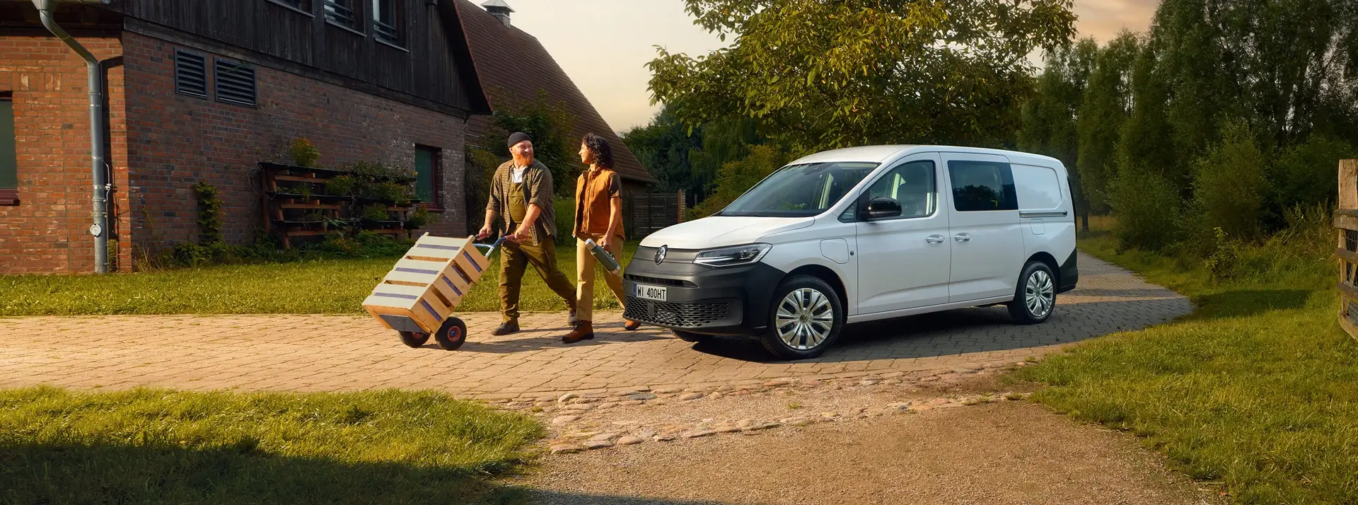 wiejskie zabudowania gospodarcze, biały Volkswagen Caddy i dwoje ludzi transportujących skrzynki niewielkim wózkiem