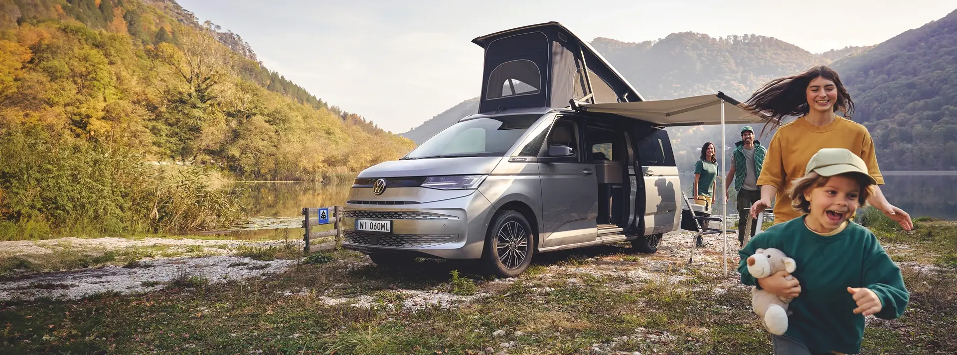 radosne dziecko i uśmiechnięta kobieta obok srebrnego Volkswagena California z rozłożonym namiotem, na łonie natury
